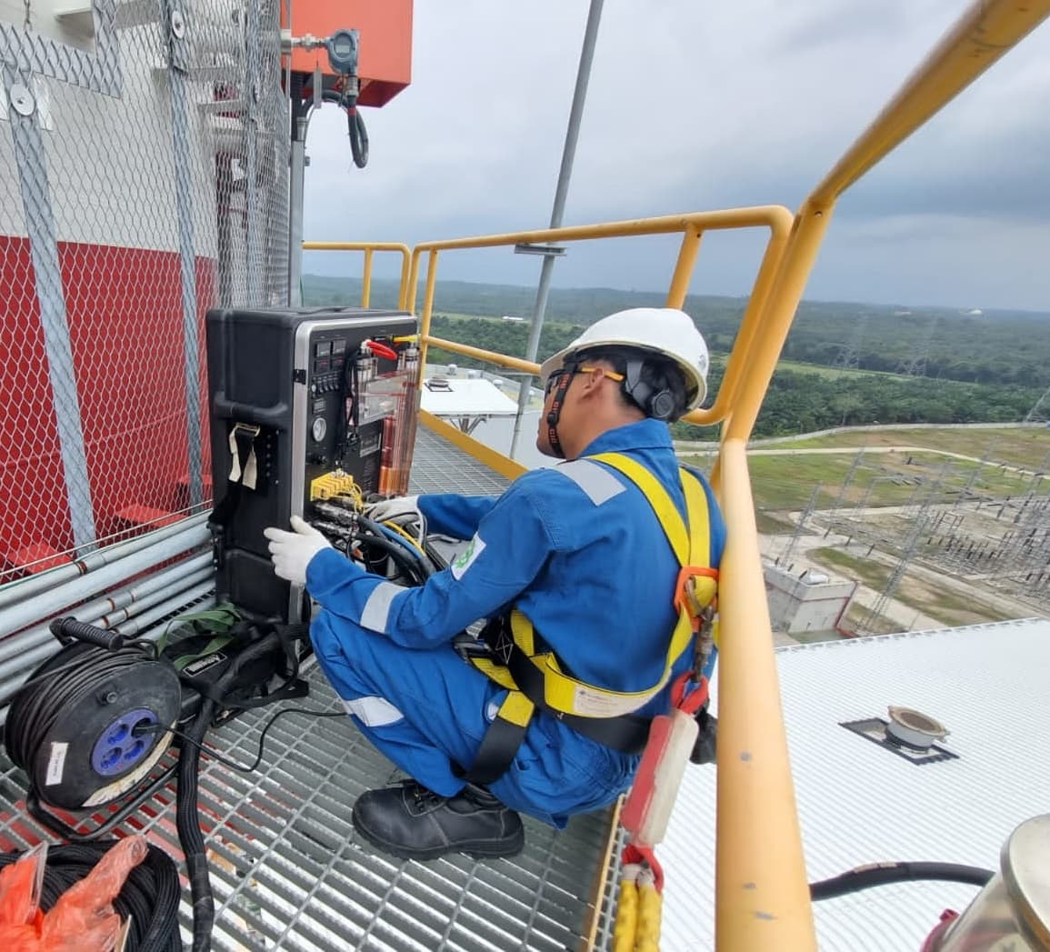 Teknisi berhelm dan harness mengoperasikan peralatan pengukuran pada platform tinggi di cerobong fasilitas industri.
