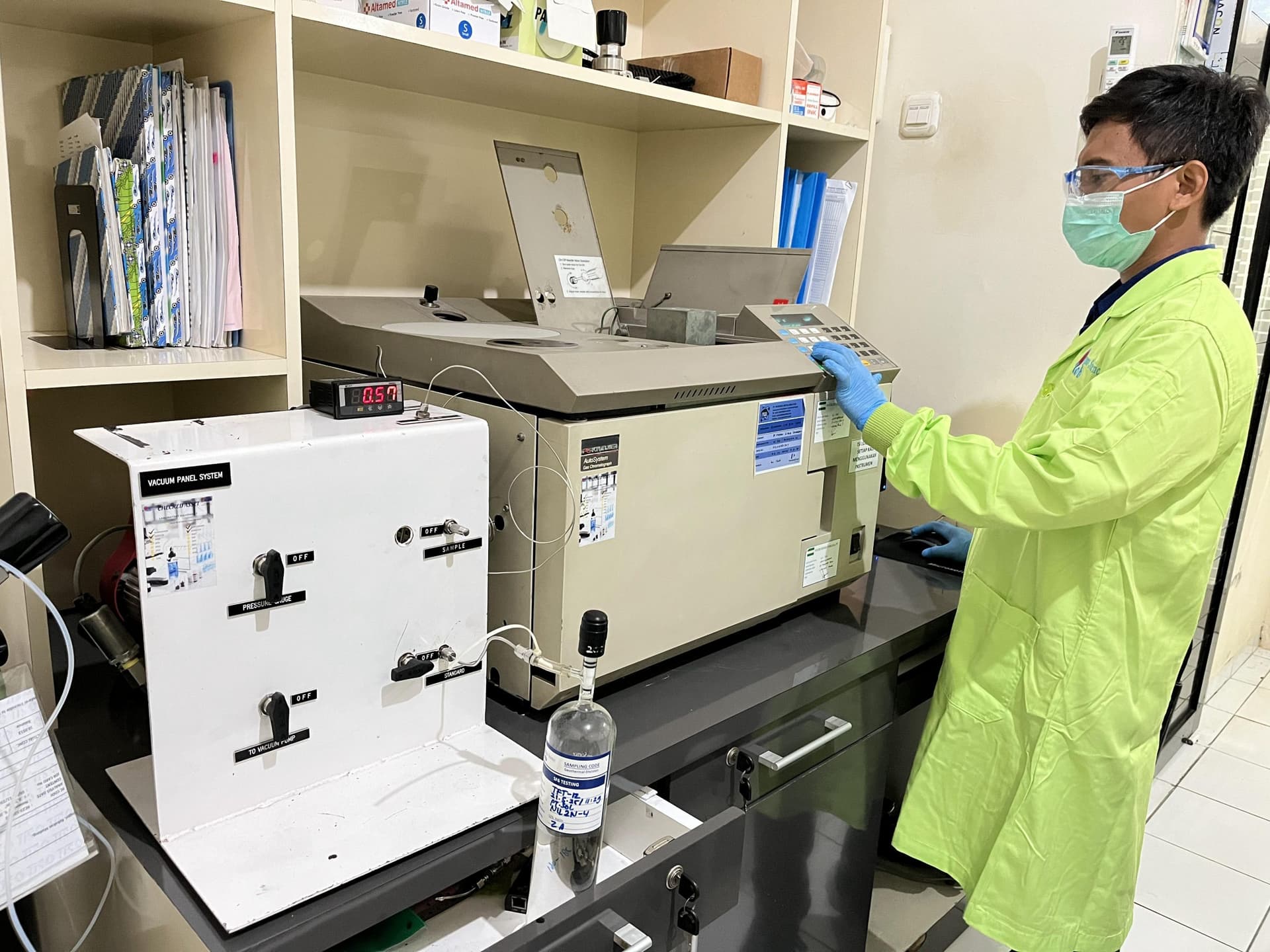 Analis berjas laboratorium hijau mengoperasikan kromatograf gas, dengan panel vakum dan botol sampel bertanda di meja.